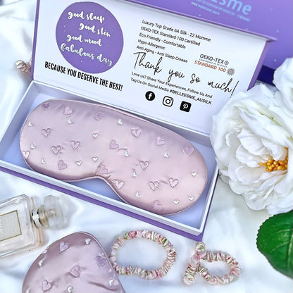A light pearl pink mulberry silk sleep mask with heart embroidery, presented in an open Belle Esme gift box